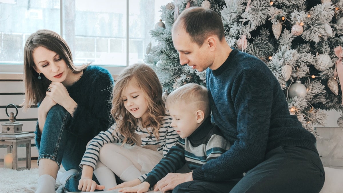 girl and boy reading book sitting between man and woman beside Christmas tree